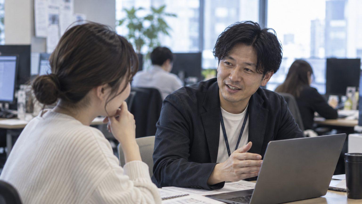オフィスの窓際でノートPCを見ながら話す2人の若手社員